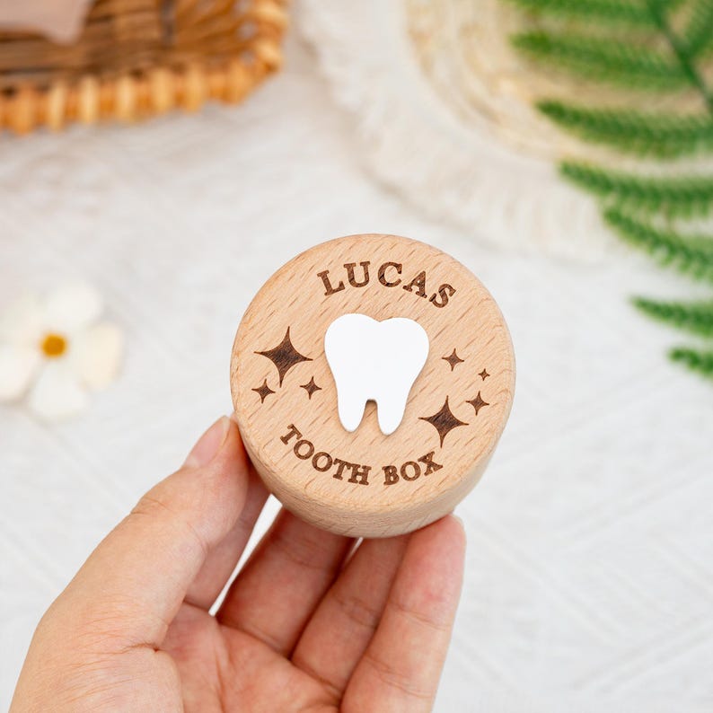 May include: A round, light-coloured wooden tooth box with a flat lid. The lid features a white tooth icon in the centre, surrounded by engraved star shapes and the text "LUCAS" at the top and "TOOTH BOX" at the bottom in a dark brown font.