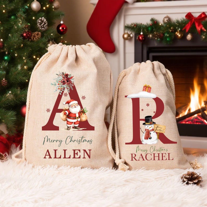 Puede incluir: Dos bolsas de regalo de arpillera beige con cord&oacute;n y dise&ntilde;os festivos. La bolsa m&aacute;s grande presenta una letra A roja, una ilustraci&oacute;n de Santa Claus y el nombre ALLEN. La bolsa m&aacute;s peque&ntilde;a presenta una letra R roja, un mu&ntilde;eco de nieve y el nombre RACHEL.