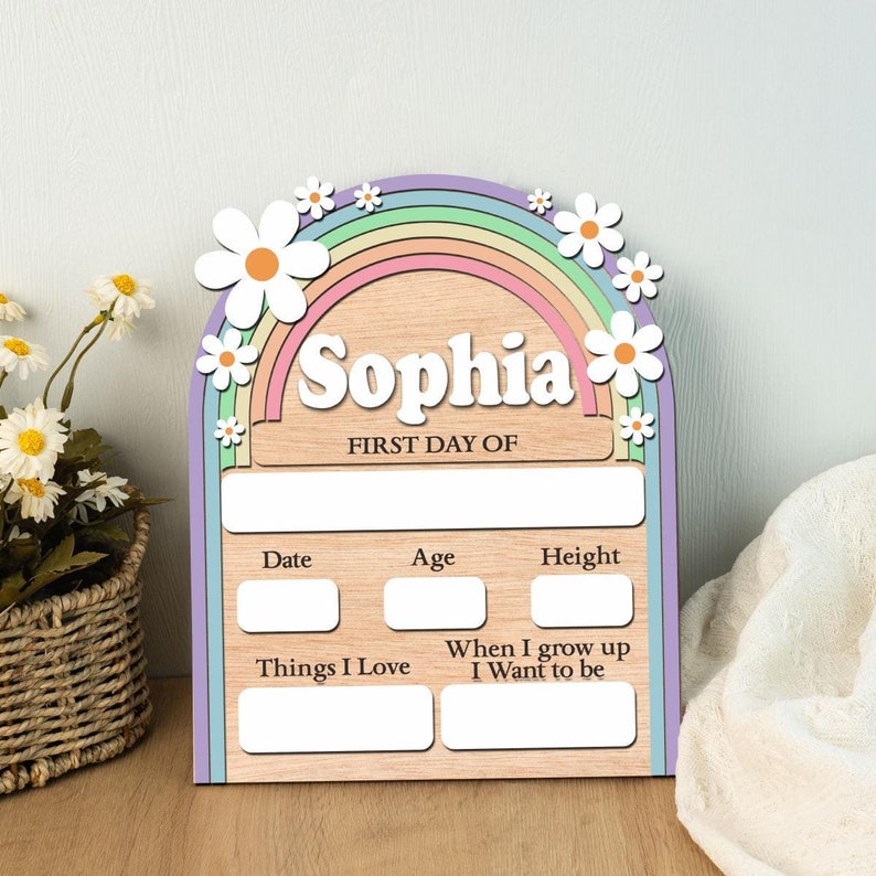 K&ouml;nnte beinhalten: Holzschild in Regenbogenform mit dem Namen "Sophia". Es hat ein pastellfarbenes Regenbogen-Design, wei&szlig;e G&auml;nsebl&uuml;mchen und Felder zur Aufzeichnung von Informationen f&uuml;r den ersten Schultag.