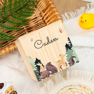 May include: A wooden box with a personalised name "Caden" and a woodland scene featuring a deer, bear, and other animals. The box is made from natural wood and has a rustic appearance.