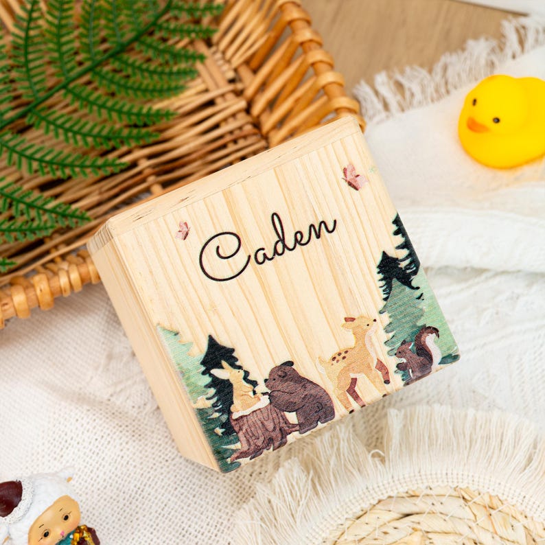 May include: A wooden box with a personalised name "Caden" and a woodland scene featuring a deer, bear, and other animals. The box is made from natural wood and has a rustic appearance.