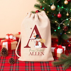 Puede incluir: Una bolsa de regalo beige con cord&oacute;n hecha de arpillera con una letra A grande de color rojo oscuro. Dentro de la letra hay un mu&ntilde;eco de nieve con sombrero negro y bufanda roja, sosteniendo un cartel que dice Merry Christmas. Debajo dice ALLEN.