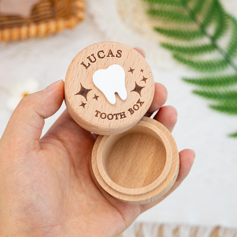 May include: A round, light-coloured wooden tooth box with a removable lid. The lid features an engraved white tooth icon in the centre, surrounded by engraved star shapes and the text "LUCAS" at the top and "TOOTH BOX" at the bottom.