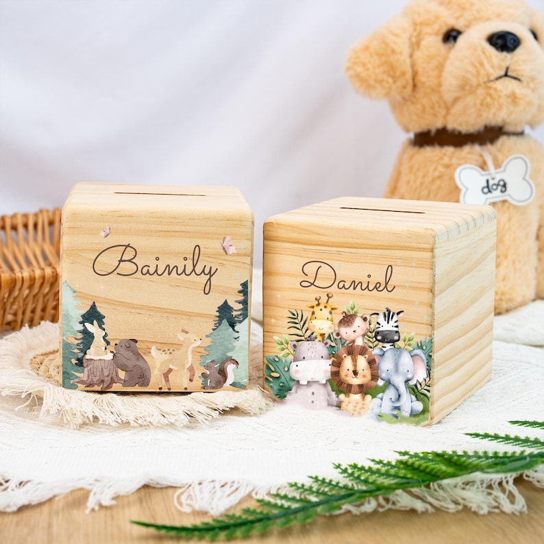 May include: Two wooden money boxes with animal illustrations. The first money box is labelled "Bainily" and features a bear, deer, rabbit, and squirrel. The second money box is labelled "Daniel" and features a giraffe, monkey, zebra, hippopotamus, lion, and elephant.