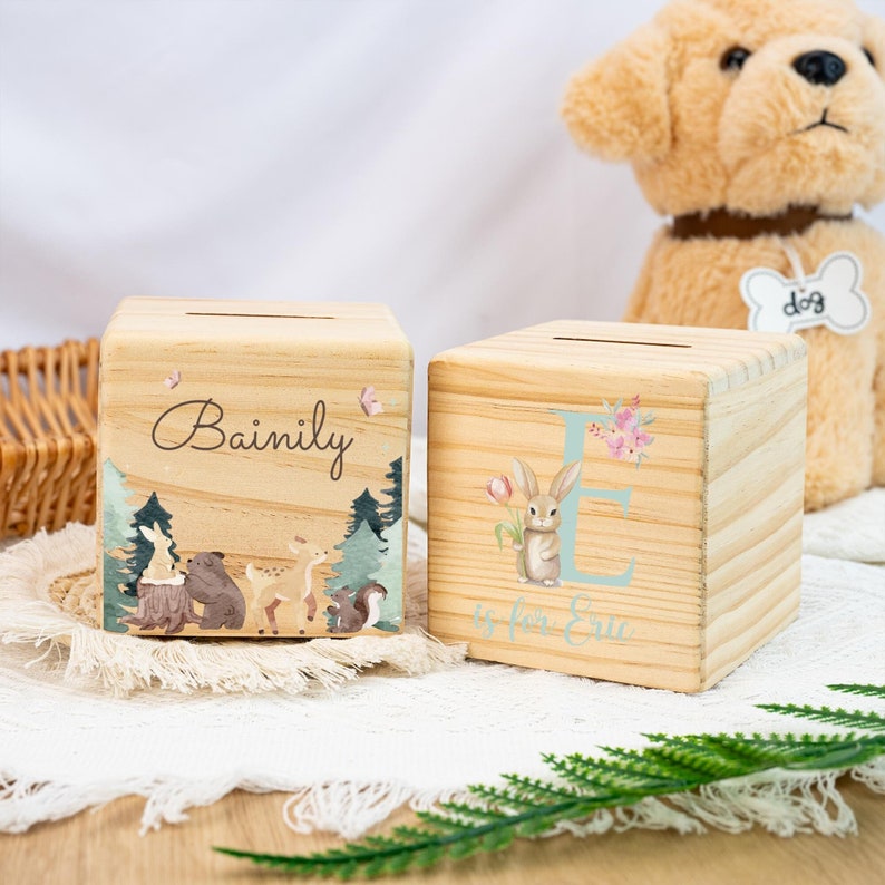 May include: Two wooden money boxes with personalised names. One box features a woodland scene with a bear, deer, and squirrel. The other box has a light blue letter E with a bunny holding a pink flower. The text "E is for Eric" is written below the letter.
