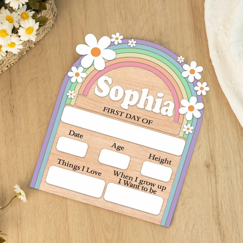 K&ouml;nnte beinhalten: Holzschild mit Regenbogen- und G&auml;nsebl&uuml;mchen-Design. Der Name "Sophia" und der Text "FIRST DAY OF" sind oben aufgedruckt. Es gibt Felder f&uuml;r Datum, Alter, Gr&ouml;&szlig;e, Interessen und Zukunftsw&uuml;nsche.