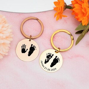 May include: Two gold keychains with round metal charms. One charm has a black handprint and footprint, the other has two black footprints and the text "James 21.06.2024".