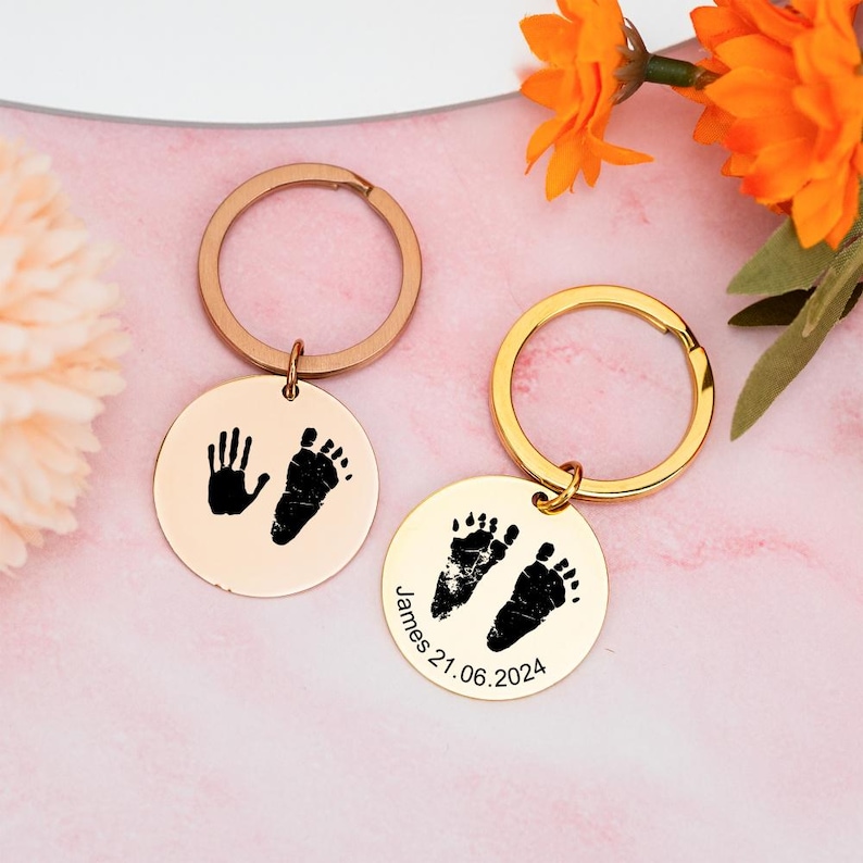 May include: Two gold keychains with round metal charms. One charm has a black handprint and footprint, the other has two black footprints and the text "James 21.06.2024".