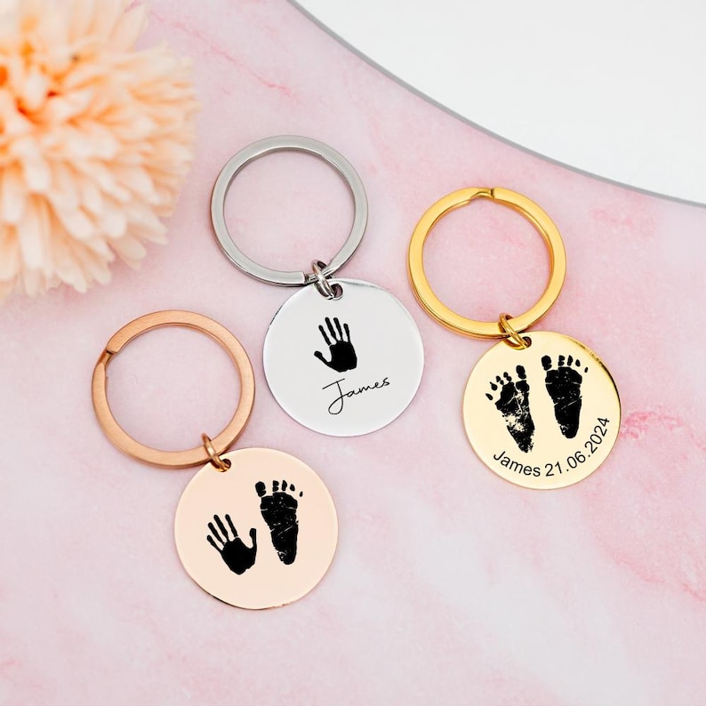 May include: Three silver keychains with round metal tags. The tags have black imprints of a handprint and footprints. The tags are engraved with the name "James" and the date "21.06.2024".