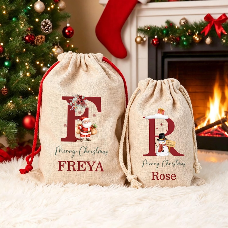 Puede incluir: Dos bolsas de regalo de arpillera beige con cord&oacute;n y gr&aacute;ficos rojos. La bolsa grande tiene una letra F roja, una ilustraci&oacute;n de Santa Claus y el nombre FREYA. La peque&ntilde;a tiene una letra R roja, un mu&ntilde;eco de nieve y el nombre ROSE.