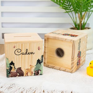 May include: Two wooden money boxes with a slot on top. One is decorated with a forest scene and the name "Caden" and the other is decorated with a red truck and the words "Jack's Donut Dreams".