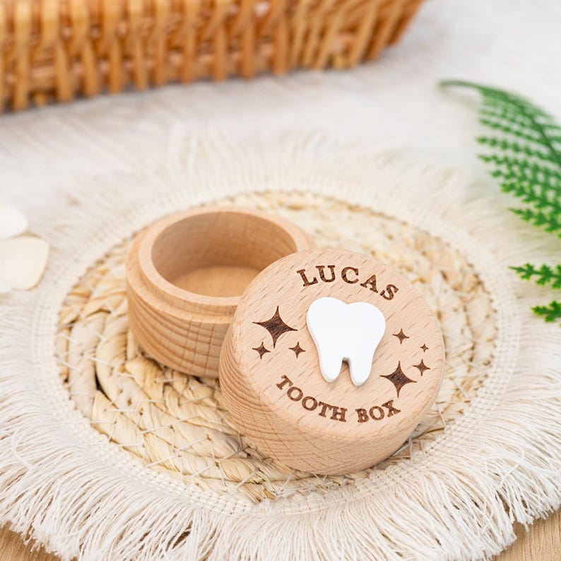 May include: A small, round, light-coloured wooden container with a removable lid. The lid features the engraved text LUCAS TOOTH BOX, a white 3D tooth icon in the centre, and several small engraved star shapes surrounding the tooth design.