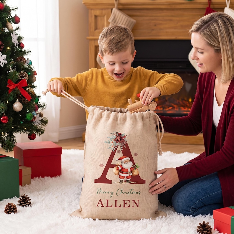 Puede incluir: Un saco de regalo de Navidad de arpillera beige con una letra A grande de color rojo oscuro, un gr&aacute;fico de Santa Claus y el texto Merry Christmas ALLEN. El saco tiene un cierre de cord&oacute;n y est&aacute; hecho de tela tejida natural.
