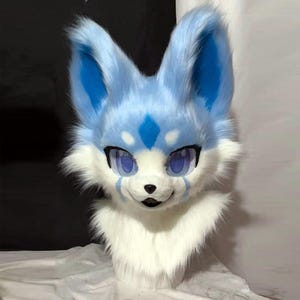 以下が含まれることがあります： 青と白の毛皮のキツネの頭。キツネは大きく表現力豊かな目を持ち、額にはひし形のマーク、耳と頬には青いアクセントがあります。毛皮は柔らかくふわふわで、遊び心のある外観を作り出しています。