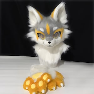 Puede incluir: Un conjunto de cabeza y patas de zorro de peluche. La cabeza es principalmente blanca, gris y amarilla, con ojos grandes y expresivos. Las patas son amarillas con almohadillas y garras blancas. El disfraz está diseñado para parecerse a un zorro de dibujos animados.