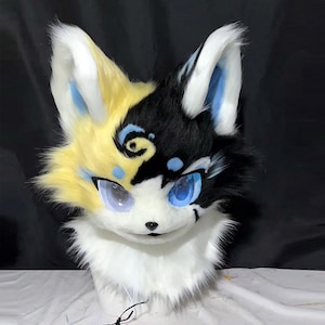May include: A furry fox head with a split design. One side is yellow and the other is black, with blue accents around the eyes and ears. The eyes are large and blue, and the muzzle is white.