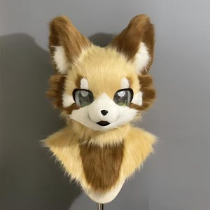 Puede incluir: Una cabeza de zorro de peluche y peluda, con grandes ojos expresivos. La máscara presenta una mezcla de pelaje marrón, beige y blanco, con rasgos faciales detallados, incluyendo una nariz negra y una boca sonriente. La cabeza de zorro está montada en un soporte.
