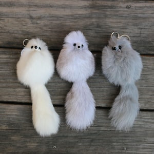 Könnte beinhalten: Drei flauschige Schlüsselanhänger mit langen Schwänzen. Die Schlüsselanhänger sind weiß, hellgrau und grau. Jeder Schlüsselanhänger hat eine kleine schwarze Knopfnase und zwei schwarze Augen.