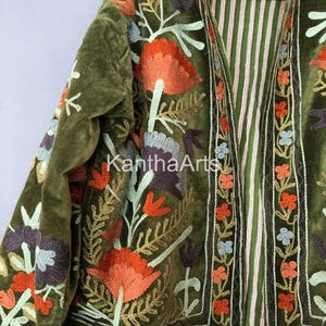 May include: An olive green velvet jacket with detailed floral embroidery in orange, purple, and blue. The jacket has a striped lining and is embellished with intricate stitching. The text "Kantha Arts" is visible.