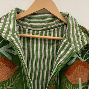May include: Green jacket with floral embroidery and a striped collar. The jacket is hanging on a wooden hanger. The embroidery features orange, blue, and green colors.