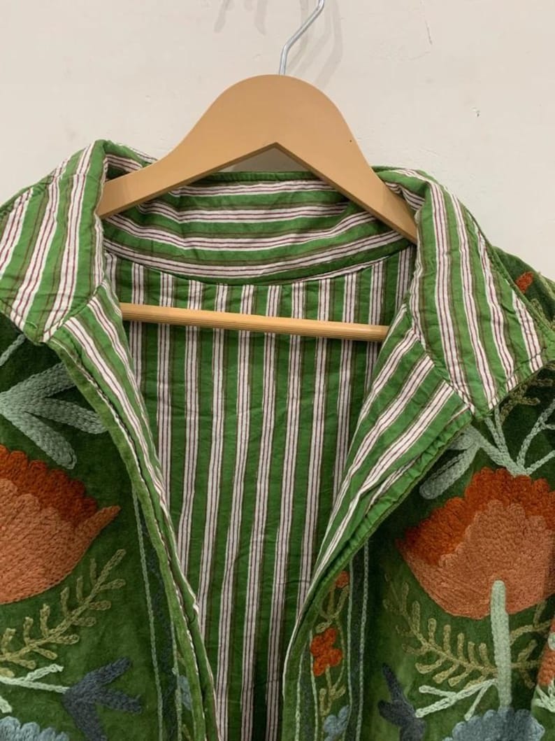 May include: Green jacket with floral embroidery and a striped collar. The jacket is hanging on a wooden hanger. The embroidery features orange, blue, and green colors.