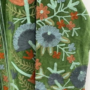 May include: Close-up of a green velvet jacket with intricate floral embroidery. The embroidery features flowers in shades of orange, blue, and green, with detailed leaf and stem designs. The jacket's texture and color create a vintage aesthetic.