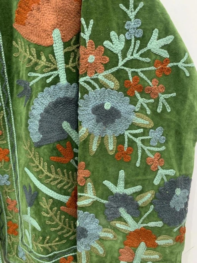 May include: Close-up of a green velvet jacket with intricate floral embroidery. The embroidery features flowers in shades of orange, blue, and green, with detailed leaf and stem designs. The jacket's texture and color create a vintage aesthetic.