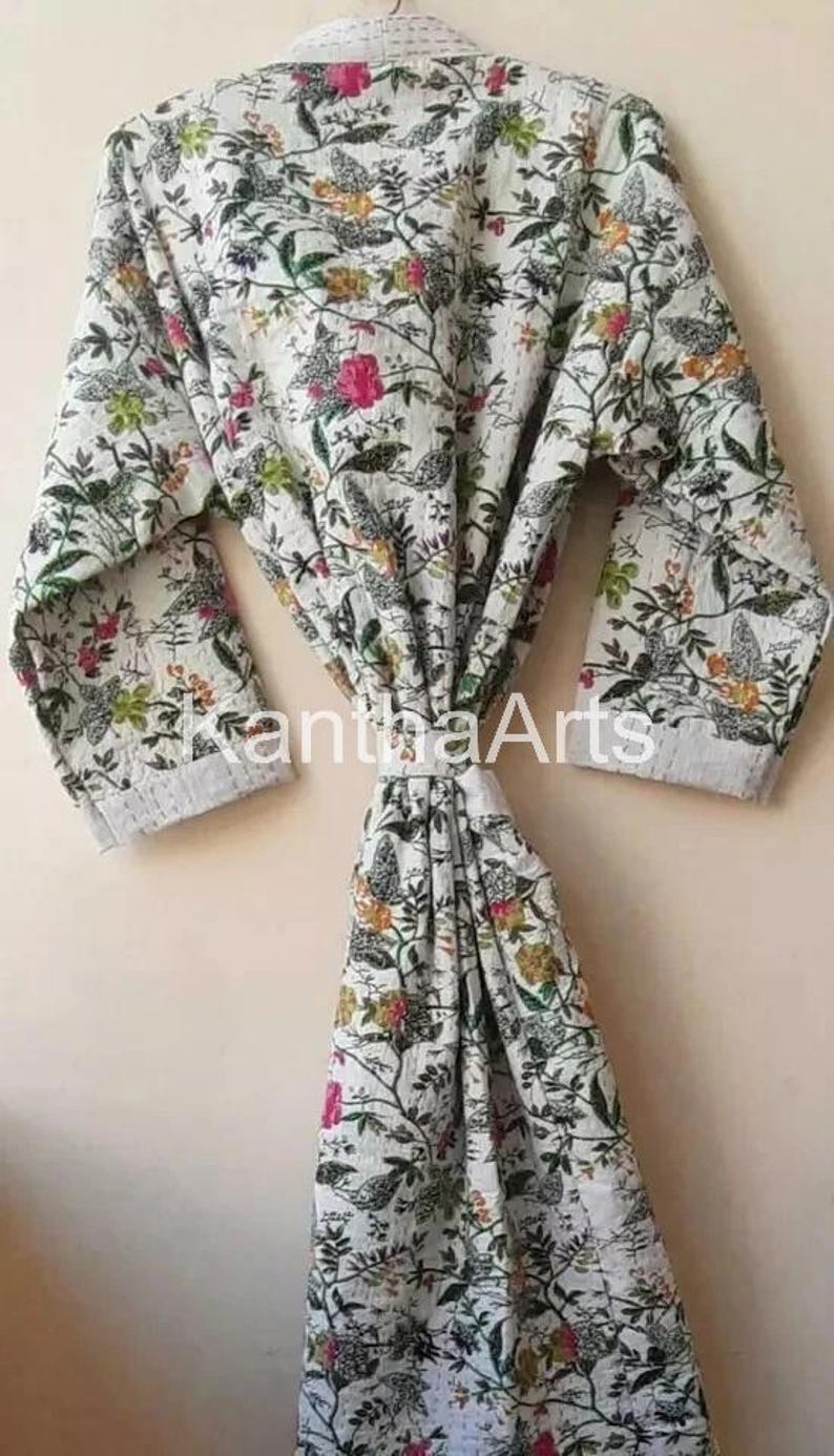 Op de afbeelding: Witte badjas met bloemenprint en een striksluiting. De badjas heeft een patroon van groene bladeren, roze en gele bloemen en vogels. De tekst "KanthaArts" is zichtbaar op de badjas.