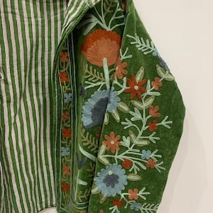 May include: Green jacket with floral embroidery on the sleeves. The jacket has a striped green and white interior. The embroidery features orange, blue, and red flowers with green leaves. The jacket is hanging on a wooden hanger.