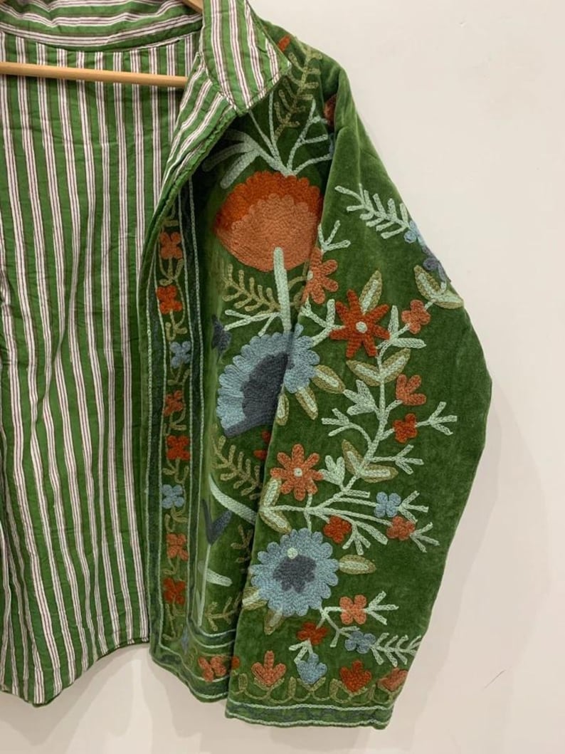 May include: Green jacket with floral embroidery on the sleeves. The jacket has a striped green and white interior. The embroidery features orange, blue, and red flowers with green leaves. The jacket is hanging on a wooden hanger.