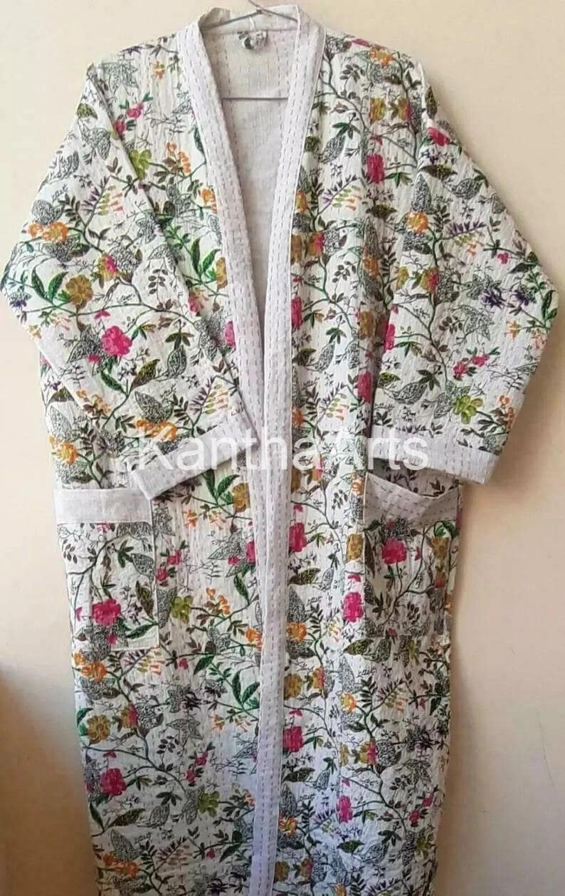 Op de afbeelding: Een witte badjas met bloemenprint, lichtgrijze afwerking en zakken. De badjas heeft een kleurrijk patroon van bloemen en bladeren in roze, groen, geel en oranje tinten. De badjas is gemaakt van een zachte, lichte stof.