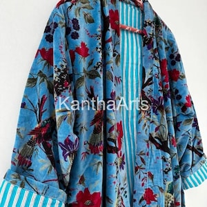 May include: A light blue velvet robe with a floral and bird pattern. The robe has red flowers and green leaves. The inside lining is striped with blue and white. The cuffs have a blue and white striped pattern. The text "Kantha Arts" is visible.