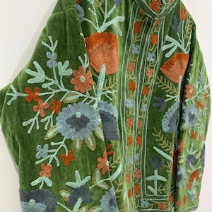 May include: A green velvet jacket with intricate floral embroidery in shades of orange, blue, and green. The jacket features a high collar and long sleeves, showcasing detailed stitching and a textured appearance. The embroidery is concentrated on the sleeves and front panels.