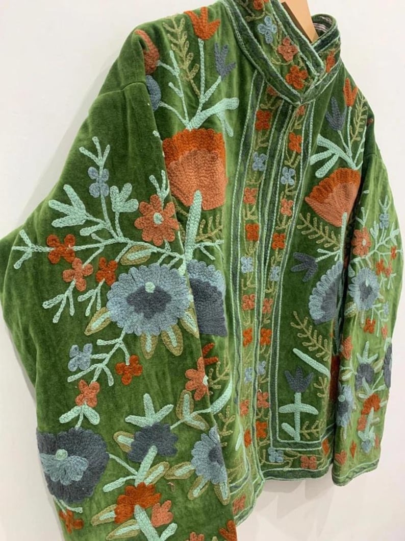 May include: A green velvet jacket with intricate floral embroidery in shades of orange, blue, and green. The jacket features a high collar and long sleeves, showcasing detailed stitching and a textured appearance. The embroidery is concentrated on the sleeves and front panels.