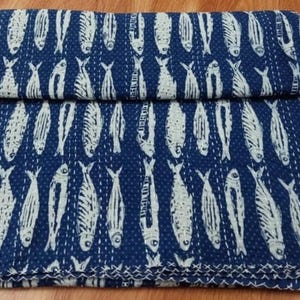 Könnte beinhalten: Eine gefaltete marineblaue Steppdecke mit einem sich wiederholenden Muster aus weißen Fischen. Die Fische sind stilisiert mit detaillierten Flossen und Schwänzen. Die Steppdecke ist aus weichem Stoff und hat einen genähten Rand.