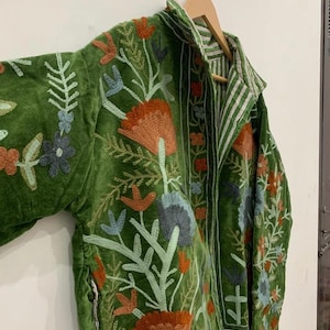 May include: Green velvet jacket with floral embroidery in orange, blue, and white. The jacket features a collared neckline and a striped interior lining. The embroidery includes detailed flower and leaf patterns.
