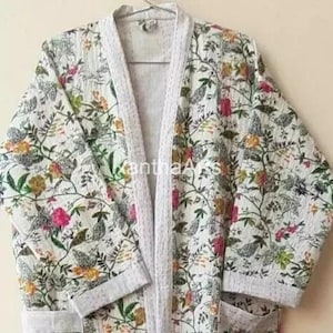 Op de afbeelding: Een witte kimono-badjas met bloemenprint en een lichtgrijze bies. De badjas heeft een kleurrijk patroon van bloemen en bladeren in roze, gele en groene tinten. De badjas heeft lange mouwen en zakken.