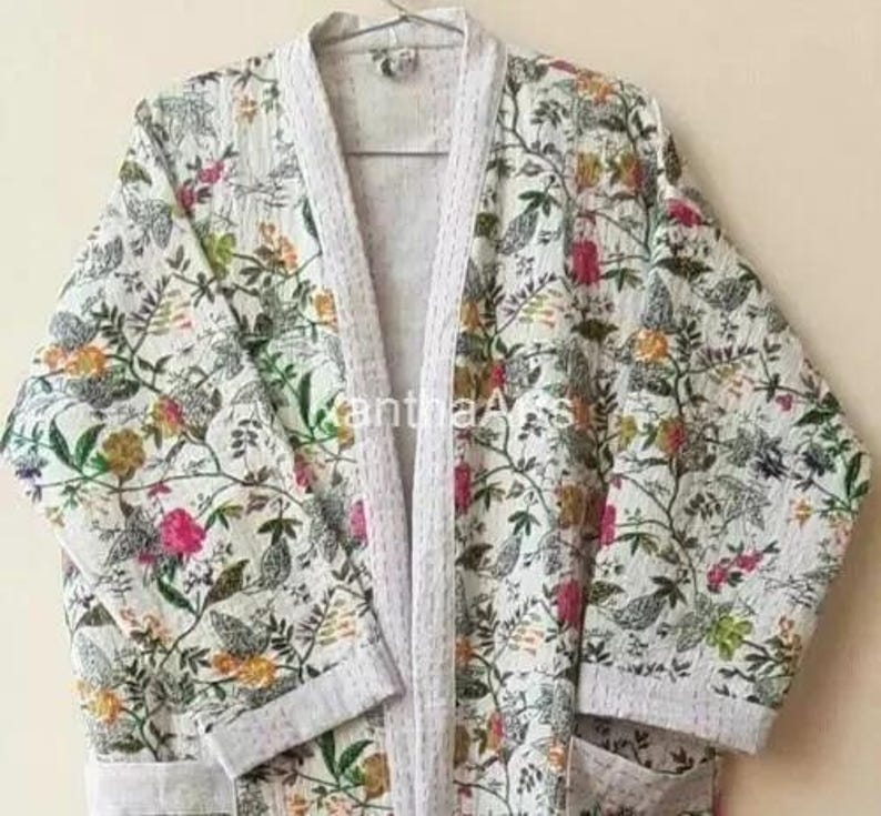 Op de afbeelding: Een witte kimono-badjas met bloemenprint en een lichtgrijze bies. De badjas heeft een kleurrijk patroon van bloemen en bladeren in roze, gele en groene tinten. De badjas heeft lange mouwen en zakken.
