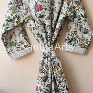 Op de afbeelding: Een witte badjas met bloemenprint en een striksluiting. De badjas heeft een patroon van groene bladeren, roze en gele bloemen en vogels. De tekst "KanthaArts" is zichtbaar op de badjas.