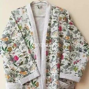 Op de afbeelding: Witte kimono-badjas met bloemenprint en lichtgrijze bies. De badjas heeft een kleurrijk patroon van bloemen en bladeren in roze, gele en groene tinten. De badjas hangt aan een metalen hanger.