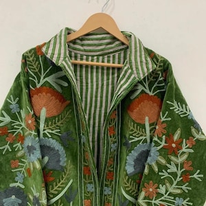 May include: Green velvet jacket with floral embroidery in orange, blue, and white. The jacket features a striped collar and lining. The embroidery is detailed and covers the front and sleeves. The jacket is hanging on a wooden hanger.