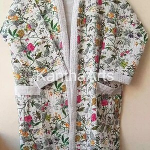 Op de afbeelding: Een witte badjas met bloemenprint, lichtgrijze afwerking en zakken. De badjas heeft een kleurrijk patroon van bloemen en bladeren in roze, groene, gele en oranje tinten. De badjas is gemaakt van een zachte, lichte stof.