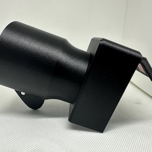 Könnte beinhalten: Ein schwarzes, zylindrisches Objekt mit strukturierter Oberfläche. Es hat ein ausgestelltes Ende und eine rechteckige Basis mit einem kleinen, abgewinkelten Ansatz. Das Objekt scheint aus Metall oder einem ähnlichen Material zu bestehen.