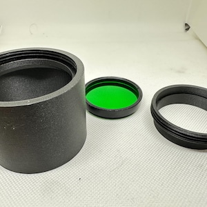 Może przedstawiać: Zestaw czarnych akcesoriów do aparatu, w tym cylindryczny adapter obiektywu, zielony filtr i pierścień gwintowany. Przedmioty są ułożone na białej powierzchni, sugerując motyw fotografii lub optyki.