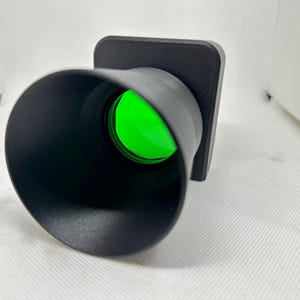 May include: A black, cylindrical device with a bright green lens. The device has a square base and a flared opening. The lens is a vibrant green color, and the device appears to be made of plastic.
