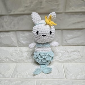 May include: A handmade crochet doll in the shape of a white bunny with a mermaid tail. The bunny has a yellow starfish on its head and is wearing a light blue and white top and a light blue mermaid tail. The doll is made of soft yarn.