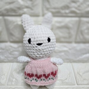 Puede incluir: Una muñeca de conejo blanca hecha a mano con ojos negros y una cara sonriente. El conejo lleva un vestido rosa con un estampado de corazones rojos y verdes. La muñeca es suave y afelpada.