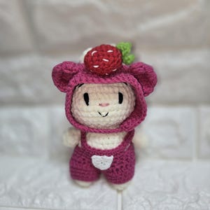 May include: A handmade crochet bunny doll wearing a pink strawberry hat and overalls. The bunny has a white body, pink accents, and a smiling face. The strawberry hat has a red berry and green leaves.