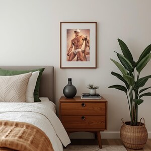 May include: A framed print of a man in a sailor's hat and briefs, leaning on a wooden ladder. The artwork is displayed above a mid-century modern nightstand with two drawers. A bed with a white comforter and a green pillow is in the foreground.