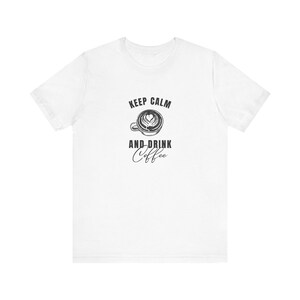 Può includere: T-shirt bianca con la scritta nera "KEEP CALM AND DRINK Coffee". Il design include una tazza di caffè con un disegno di latte art a forma di cuore. Un capo casual.